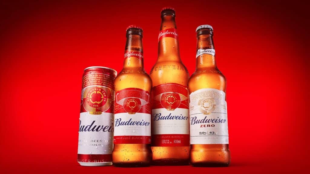 Quatro garrafas de cerveja Budweiser, incluindo rótulos temáticos da Copa do Mundo de Clubes da FIFA 2025, sobre fundo vermelho.