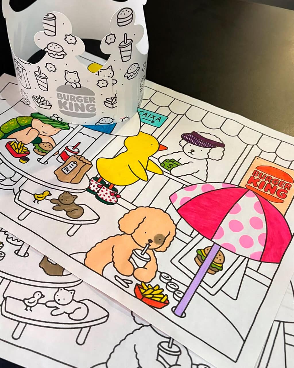 Uma coroa de papel do Burger King ao lado de desenhos para colorir mostrando uma cena de restaurante com animais divertidos, incluindo um pato e um cachorro. Os desenhos apresentam elementos como sanduíches, fritas e uma sombrinha.