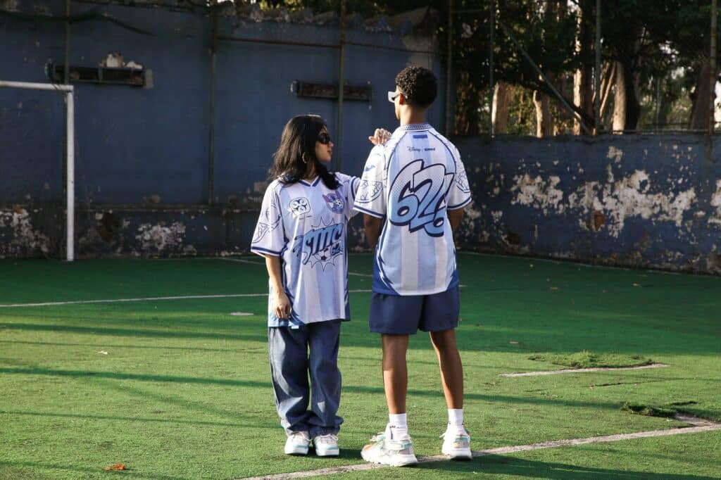 Duas pessoas usando camisetas jersey exclusivas da colaboração entre Kings e Lilo & Stitch, em um campo de futebol com grama sintética. Um jovem com camiseta azul e branca está virado de costas, enquanto uma jovem de cabelo longo com camiseta semelhante está de frente, ambas posando de forma descontraída.