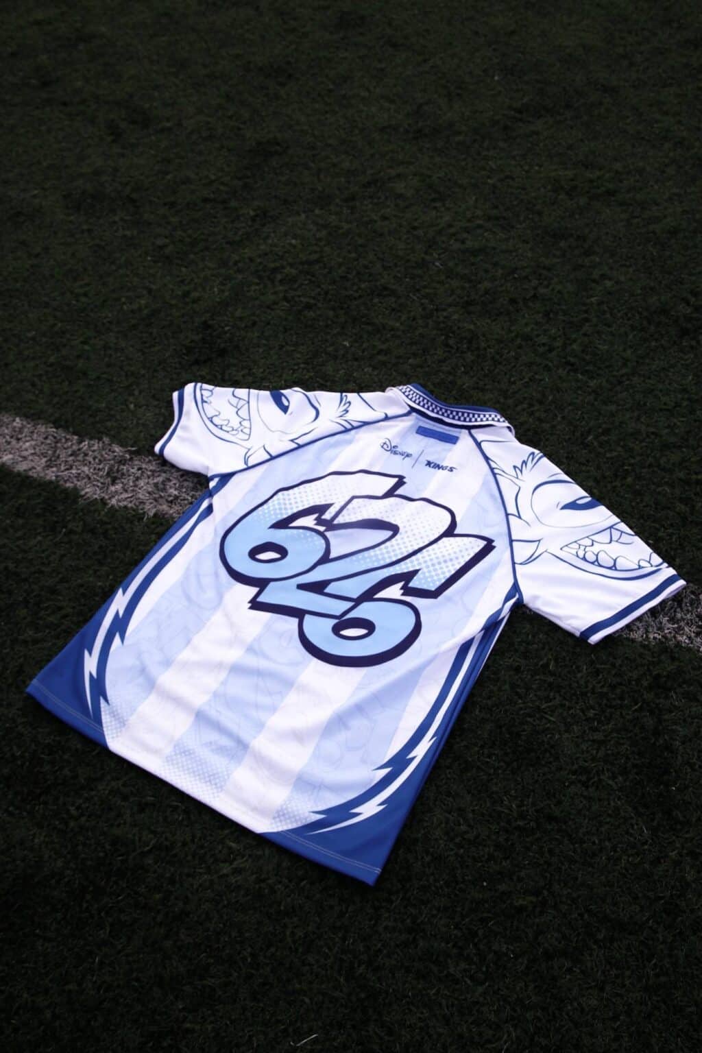 Camiseta jersey branca e azul com estampa do Stitch e o número 62, sobre um campo de grama sintética.