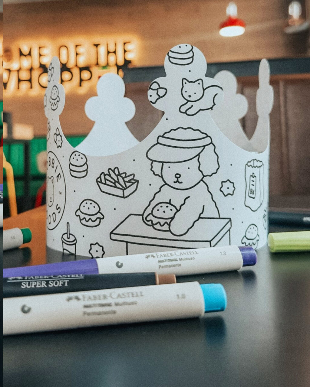 Coroa de papel em branco com desenhos para colorir, incluindo um animal e fast food, cercada por canetinhas Faber-Castell sobre uma mesa.
