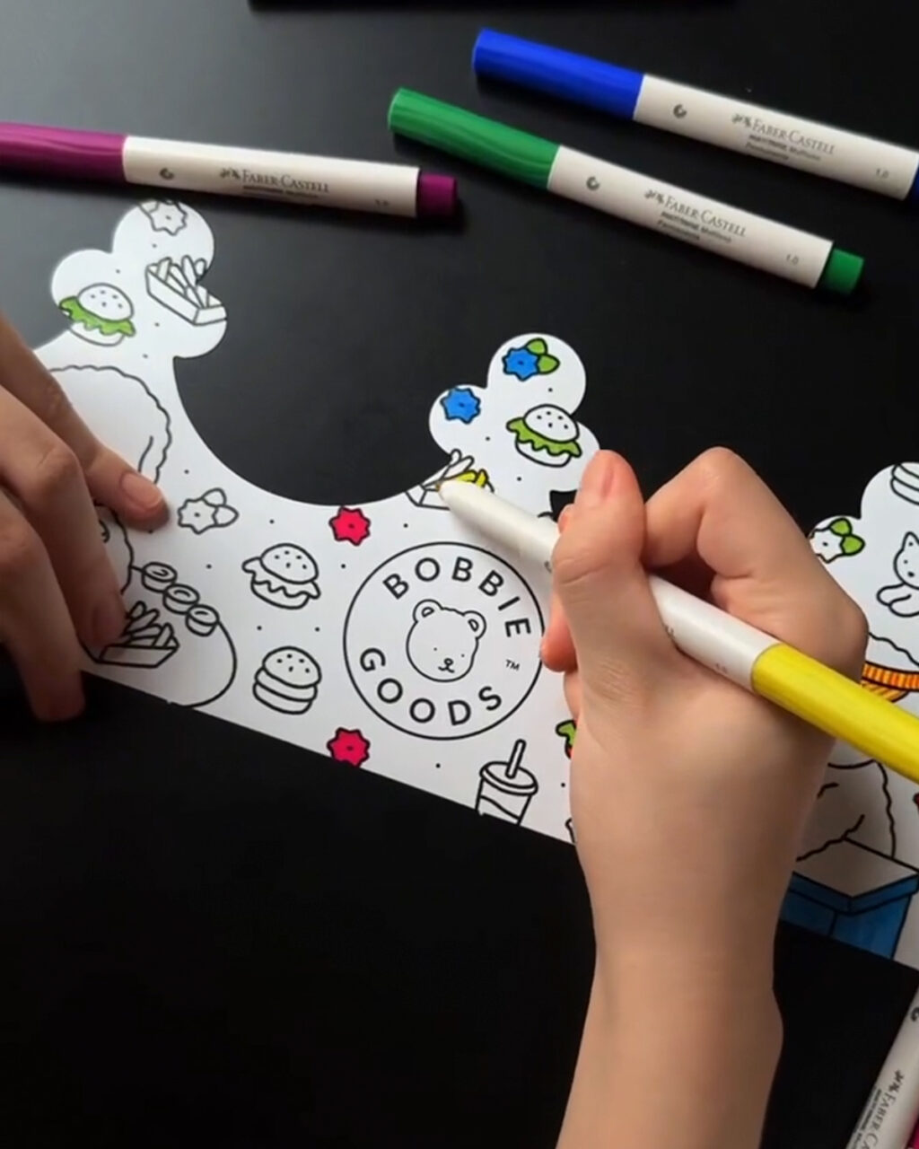 Mãos colorindo uma coroa de papel com desenhos diversos, incluindo lanches e o logo Bobbie Goods, acompanhadas de canetinhas da Faber-Castell sobre uma mesa.