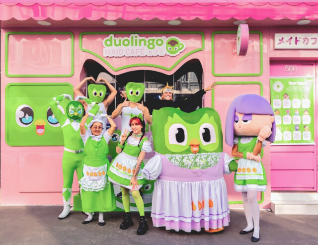 Grupo de pessoas vestindo roupas verdes e fantasias do Duolingo em frente ao Duolingo Maid Café, um estabelecimento colorido em estilo japonês.