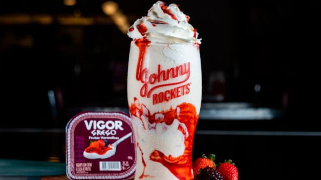 Milkshake da Johnny Rockets servido em copo alto com calda e chantilly, feito com iogurte grego Vigor de frutas vermelhas, ao lado do pote do iogurte e frutas frescas como morango e amora.