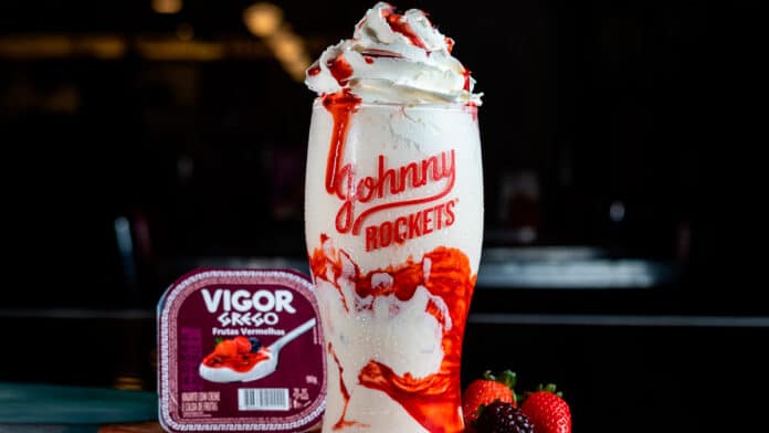 Milkshake da Johnny Rockets servido em copo alto com calda e chantilly, feito com iogurte grego Vigor de frutas vermelhas, ao lado do pote do iogurte e frutas frescas como morango e amora.