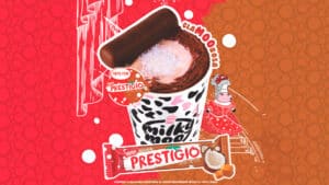 Copo da Milky Moo com estampa de vaca, recheado com sobremesa de chocolate, coco e doce de leite, decorado com pedaço de chocolate Prestígio. Ao fundo, ilustração colorida com vaca estilizada e embalagem da barra Prestígio edição limitada sabor doce de leite. Imagem promocional da parceria Milky Moo e Nestlé.
