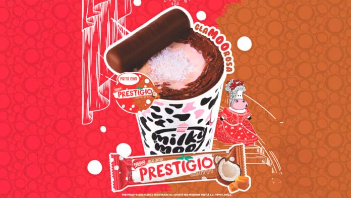 Copo da Milky Moo com estampa de vaca, recheado com sobremesa de chocolate, coco e doce de leite, decorado com pedaço de chocolate Prestígio. Ao fundo, ilustração colorida com vaca estilizada e embalagem da barra Prestígio edição limitada sabor doce de leite. Imagem promocional da parceria Milky Moo e Nestlé.