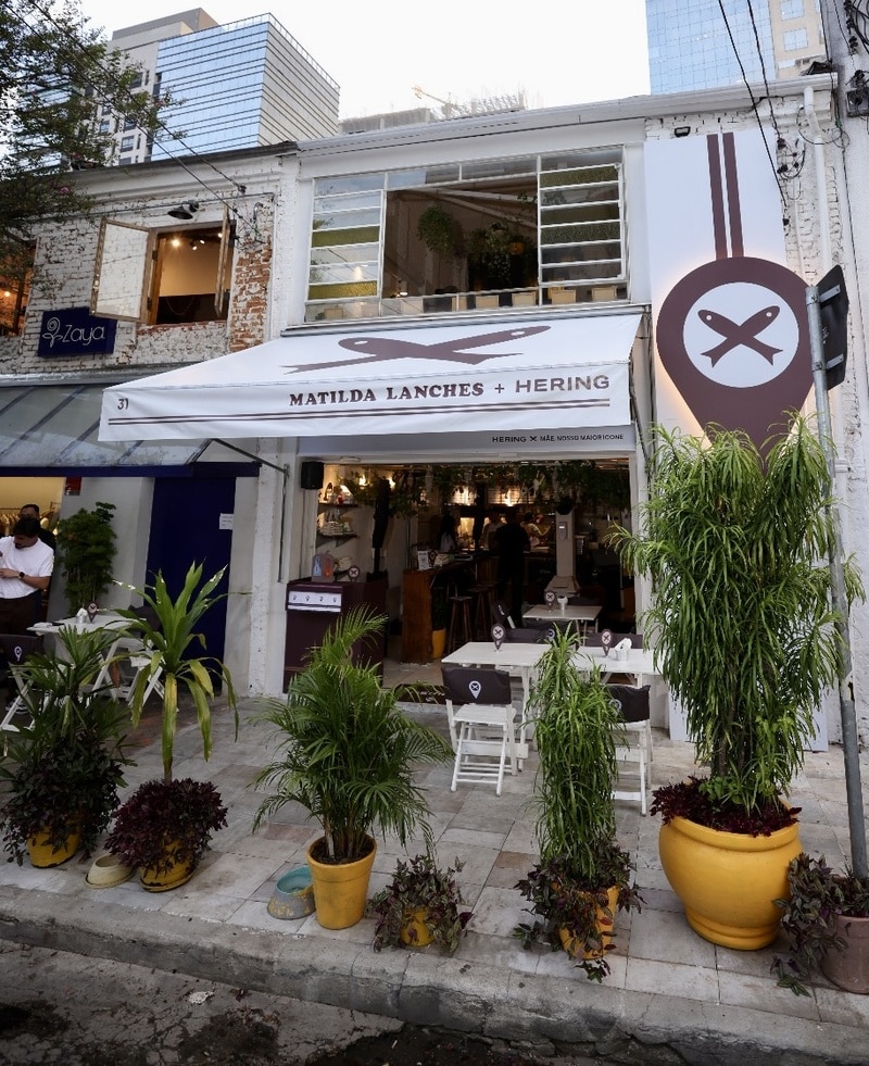 Fachada do restaurante Matilda Lanches com a marca Hering em Pinheiros, São Paulo. A entrada é decorada com plantas em vasos coloridos e mesas brancas dispostas na calçada.