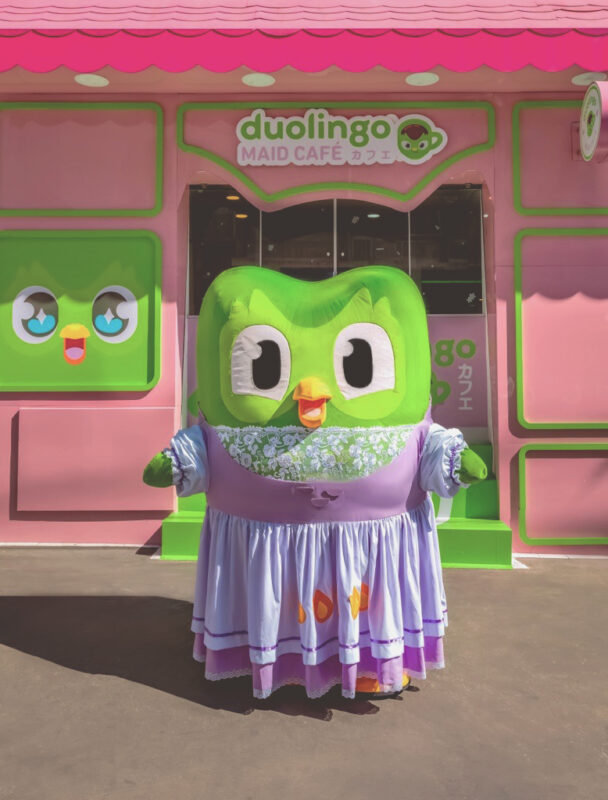 Uma figura do Duolingo vestida com um traje de maid, posando em frente ao café temático Duolingo Maid Café, que tem uma decoração em tons de rosa e verde.