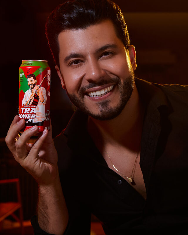 Homem sorrindo, segurando uma lata da bebida energética Extra Power, com design exclusivo do cantor Murilo Huff. Ele usa uma camisa preta e exibe um colar, em um ambiente iluminado.