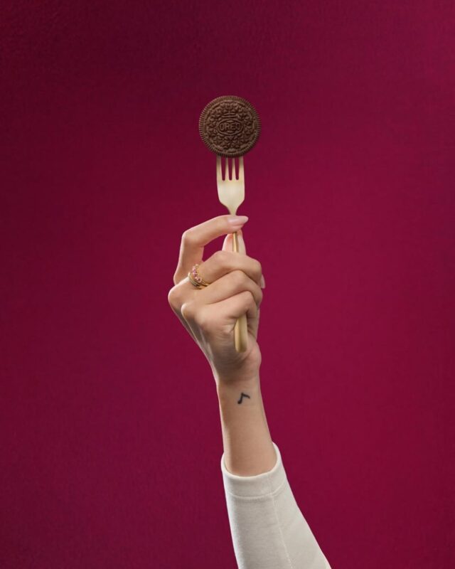Mão segurando um biscoito Oreo em um garfo, com um fundo de cor sólida.