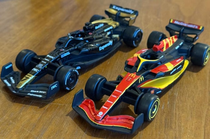 Duas miniaturas de carros de Formula 1, uma em preto e dourado e outra nas cores vermelho e amarelo, dispostas sobre uma superfície de madeira.