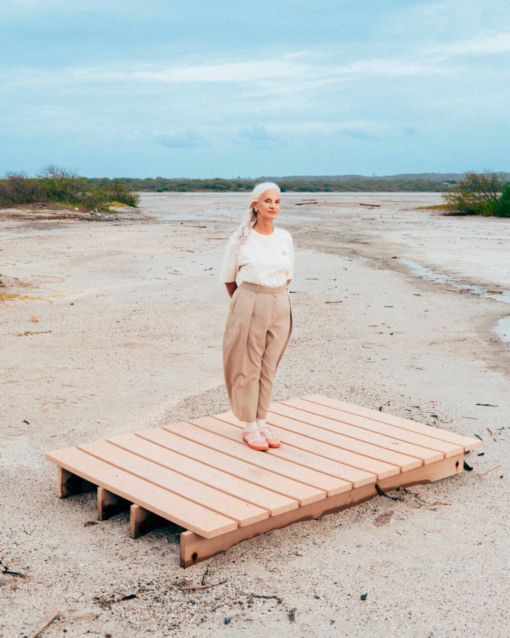 Uma mulher idosa está de pé sobre uma plataforma de madeira em um ambiente de praia, com fundo de céu azul e vegetação ao longe. Ela veste uma camiseta clara e calças bege, com um par de tênis rosa.