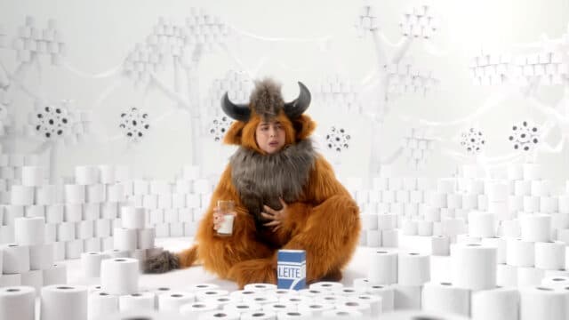 Um personagem vestido com um traje de mamífero e chifres segura um copo de leite enquanto está cercado por rolos de papel higiênico em um cenário branco.