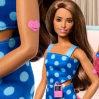 Boneca Barbie com diabetes tipo 1 vestindo um vestido azul de bolinhas, com um sensor contínuo de glicose no braço e uma bomba de insulina na cintura.