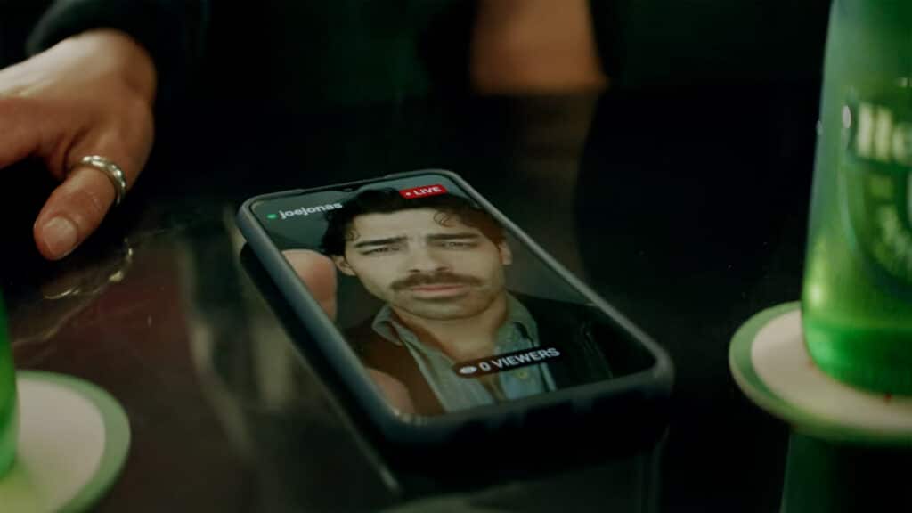gkpb-joe-jonas-heineken Celular sobre uma mesa exibindo uma live do cantor Joe Jonas no Instagram, com a indicação "LIVE" e o nome de usuário "@joejonas". A transmissão mostra Joe Jonas com expressão séria, bigode e cabelo ondulado. A live está com "0 viewers". Ao redor do celular, é possível ver parte da mão de uma pessoa com anel prateado e duas garrafas verdes da marca Heineken sobre porta-copos. A imagem sugere um ambiente de bar ou restaurante.