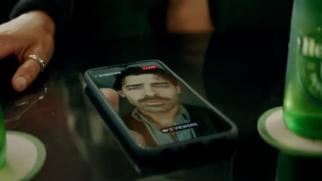 Celular sobre uma mesa exibindo uma live do cantor Joe Jonas no Instagram, com a indicação "LIVE" e o nome de usuário "@joejonas". A transmissão mostra Joe Jonas com expressão séria, bigode e cabelo ondulado. A live está com "0 viewers". Ao redor do celular, é possível ver parte da mão de uma pessoa com anel prateado e duas garrafas verdes da marca Heineken sobre porta-copos. A imagem sugere um ambiente de bar ou restaurante.