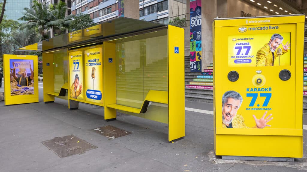 gkpb-karaoke-mercado-livre Ponto de ônibus amarelo temático de karaokê do Mercado Livre na Avenida Paulista, com publicidade sobre a campanha de descontos 7.7 e uma tela interativa para cantar e liberar cupons.