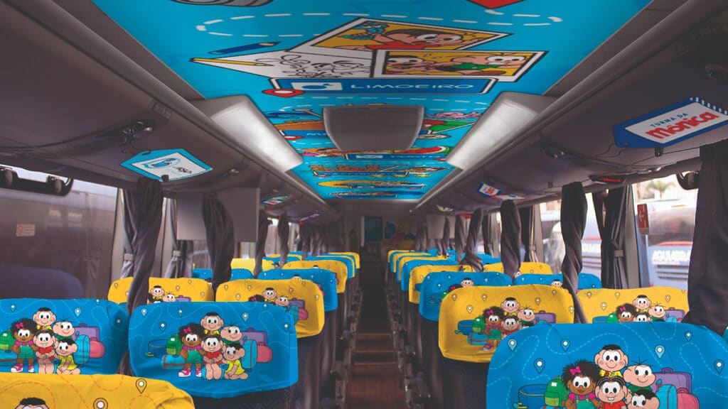 Interior do ônibus temático da Turma da Mônica, com assentos decorados e teto colorido, apresentando os personagens e temática infantil.