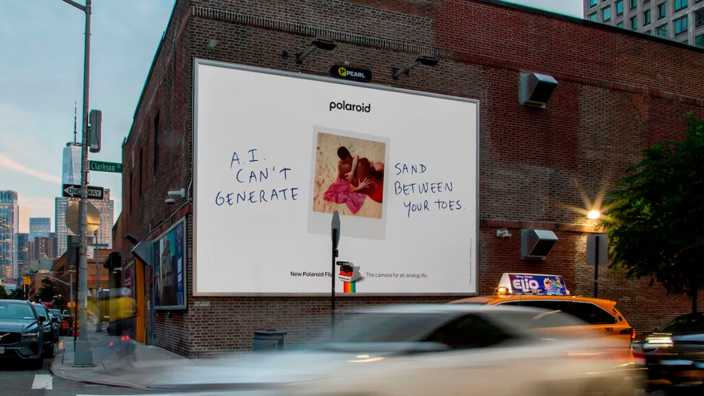 Propaganda externa em Outdoor da campanha da Polaroid com o texto "I.A. NÃO PODE GERAR AREIA ENTRE OS DEDOS DOS SEUS PÉS" ao lado da imagem de uma pessoa com uma câmera Polaroid.