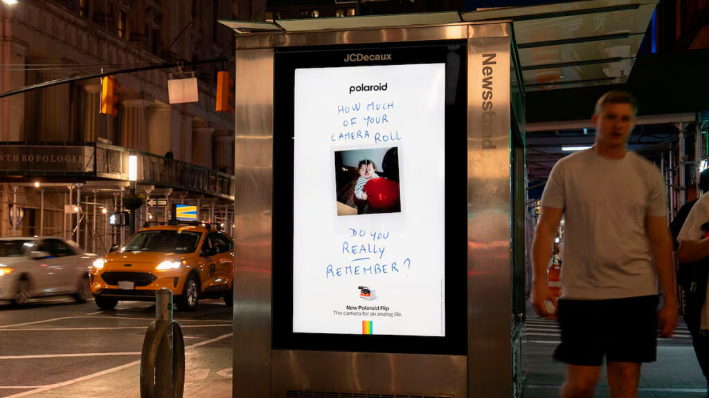 Um outdoor da Polaroid com uma foto de uma criança segurando um balão vermelho, acompanhada de anotações manuscritas que questionam sobre quantos momentos da sua galeria do celular você realmente se lembra.