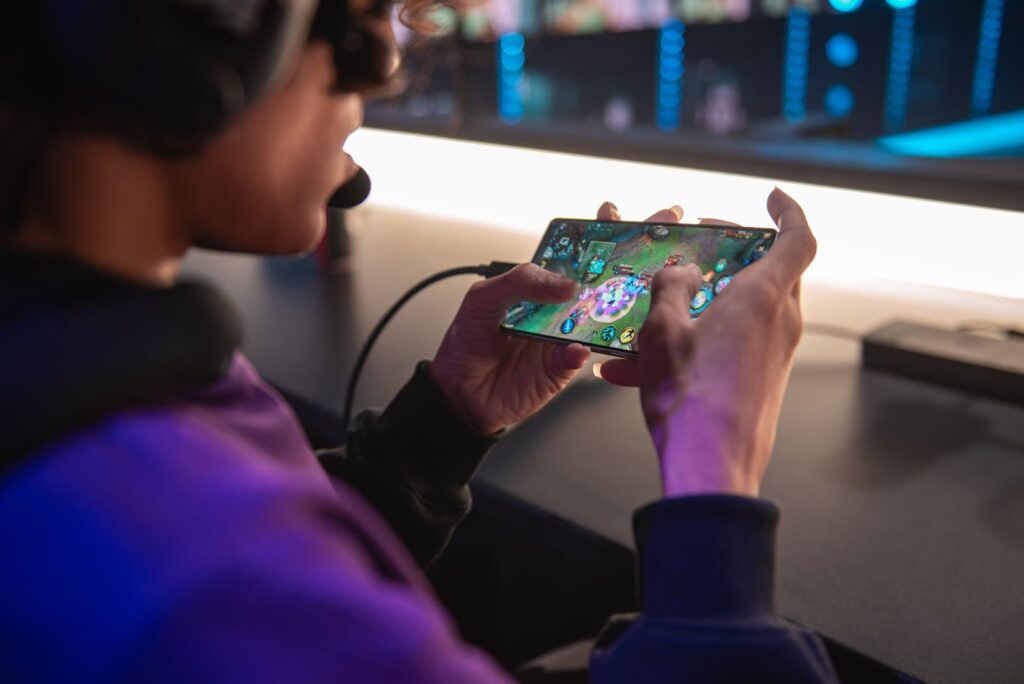 Jogador utilizando um smartphone para jogar Wild Rift em um ambiente de competição, com foco nas mãos segurando o dispositivo e a tela do jogo visível.