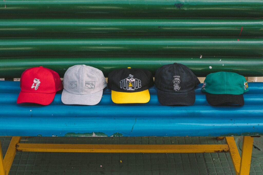 Cinco bonés da coleção Tupode da Turma da Mônica, dispostos em uma bancada azul, com cores e bordados variados, incluindo um boné vermelho, um cinza, um preto com detalhes amarelos, um preto e um verde.