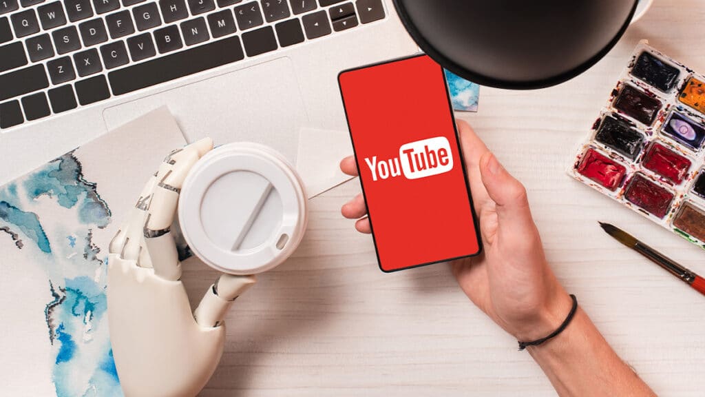 Uma mão humana segurando um smartphone com o logo do YouTube, ao lado de uma mão robótica segurando um copo. O fundo contém um laptop, uma lâmpada e materiais de pintura.
