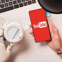 Uma mão humana segurando um smartphone com o logo do YouTube, ao lado de uma mão robótica segurando um copo. O fundo contém um laptop, uma lâmpada e materiais de pintura.