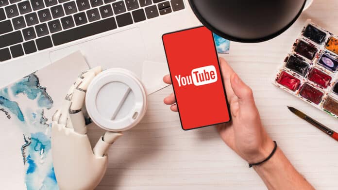 Uma mão humana segurando um smartphone com o logo do YouTube, ao lado de uma mão robótica segurando um copo. O fundo contém um laptop, uma lâmpada e materiais de pintura.