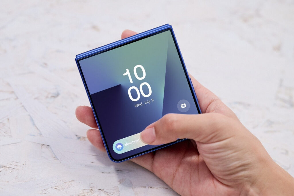 Mão segurando o smartphone dobrável Galaxy Z Flip7 com a tela externa visível, mostrando a hora e data.