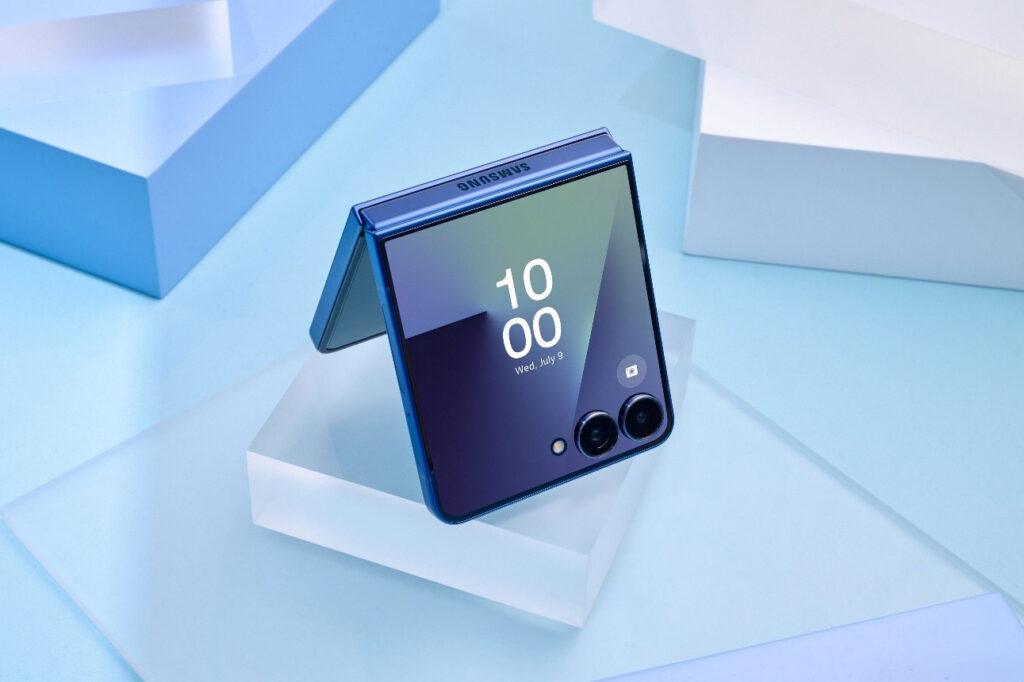 O Samsung Galaxy Z Flip7 aberto, exibindo uma tela interna com hora e data, em um fundo colorido com formas geométricas.