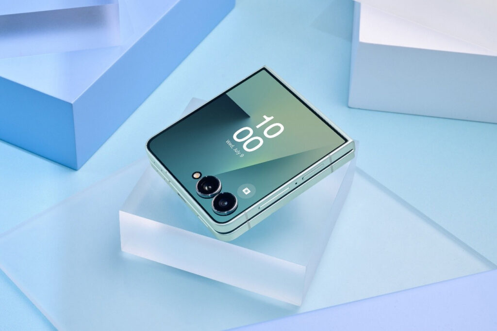 Smartphone dobrável Galaxy Z Flip7 em um suporte transparente sobre uma superfície azul, exibindo a tela com relógio e data.