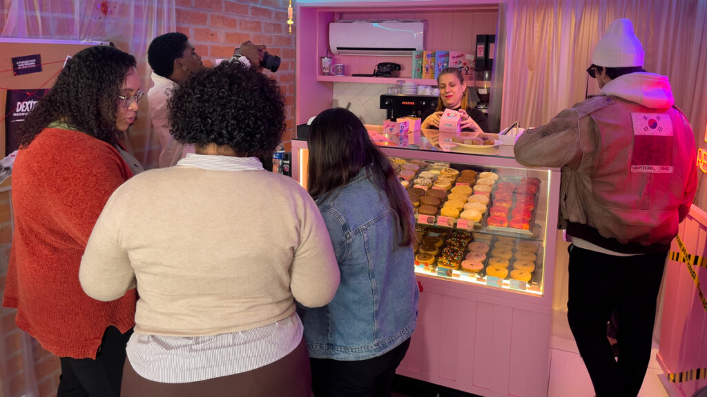 Clientes na loja Good Cop Donut interagindo com a funcionária enquanto apreciam uma vitrine repleta de donuts coloridos, em um ambiente temático inspirado na série Dexter.