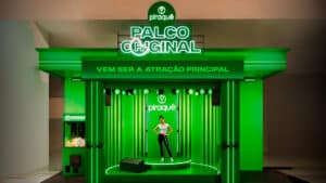 Estande verde da Piraquê chamado 'Palco Original', com iluminação de LED e a frase 'Vem ser a atração principal' em destaque. No centro, há um palco iluminado onde uma mulher posa em frente ao logotipo da marca. À esquerda, uma máquina de prêmios personalizada com a identidade visual da Piraquê.