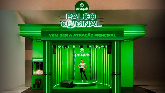 Estande verde da Piraquê chamado 'Palco Original', com iluminação de LED e a frase 'Vem ser a atração principal' em destaque. No centro, há um palco iluminado onde uma mulher posa em frente ao logotipo da marca. À esquerda, uma máquina de prêmios personalizada com a identidade visual da Piraquê.