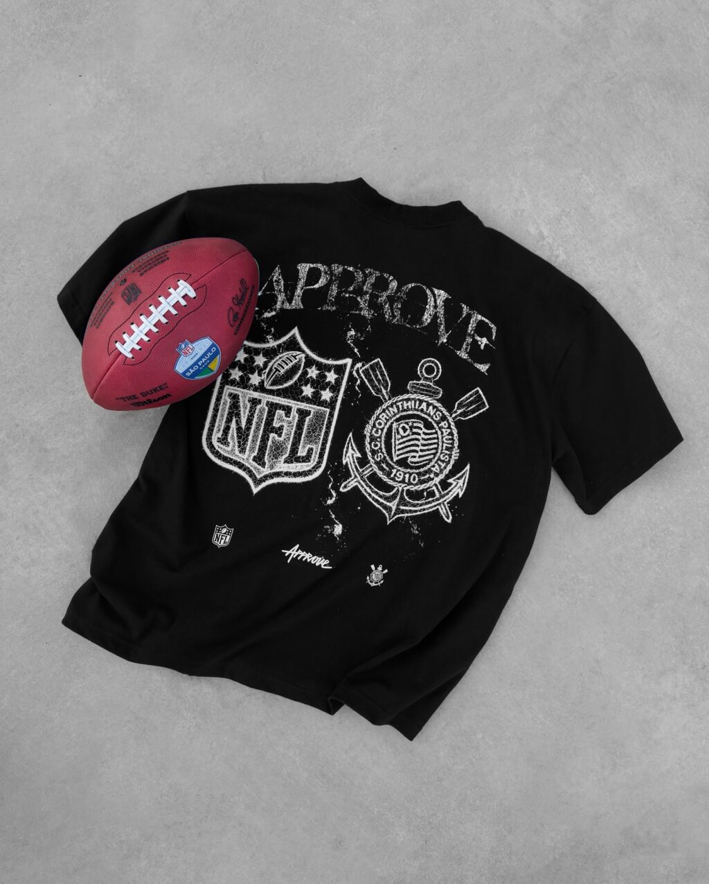 Camiseta preta da coleção entre Corinthians e NFL com o logo da NFL e do Corinthians estampado na parte de trás, ao lado de uma bola de futebol americano.