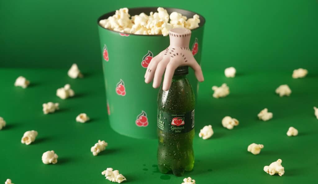 Garrafa de Guaraná Antarctica com uma miniatura da mão segurando a tampa, ao lado de um balde de pipoca sobre um fundo verde.