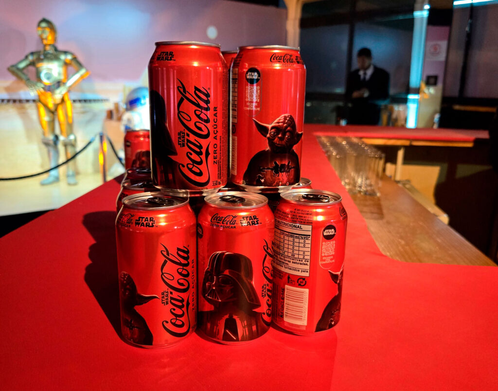 Latas de Coca-Cola edição especial Star Wars empilhadas em uma mesa vermelha, com personagens icônicos como Yoda e Darth Vader ilustrados nas embalagens. Ao fundo, personagens de Star Wars podem ser vistos em uma projeção.