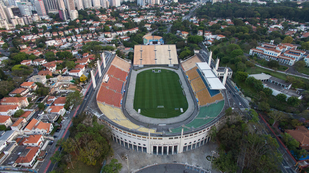 Vista aérea do Estádio Pacaembu, cercado por edifícios e áreas residenciais, mostrando o gramado e as arquibancadas.