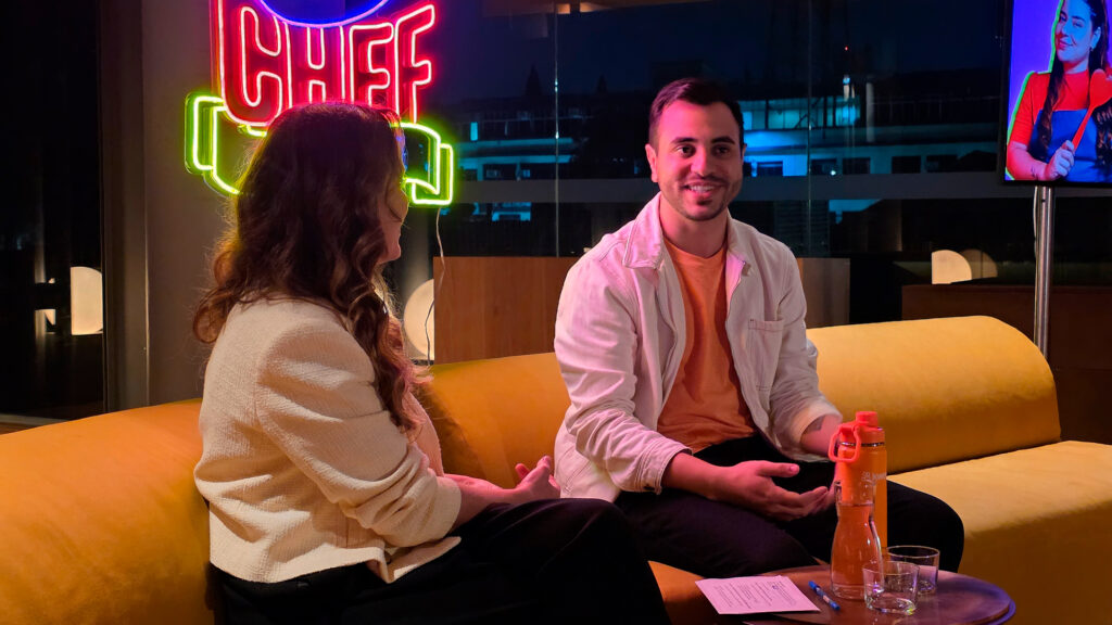 Apresentadores sentados em um sofá com uma iluminação suave, discutindo sobre o programa Kwai Chef, ao fundo uma placa de néon do programa.