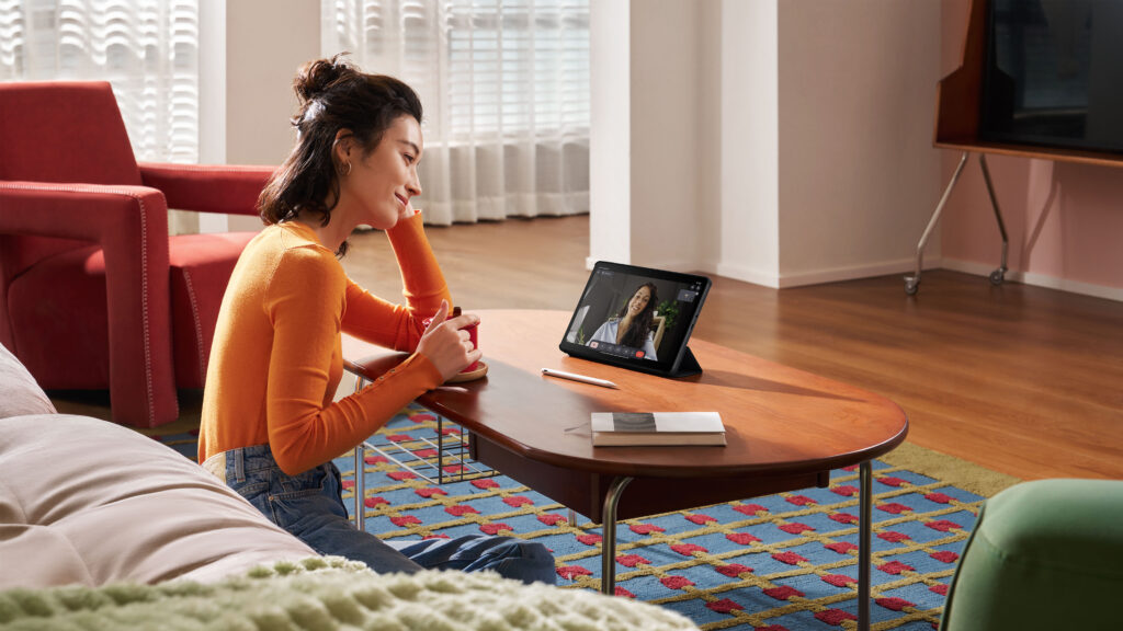 Mulher usando um tablet em uma mesa de café, assistindo a uma videochamada, em uma sala bem iluminada com decoração moderna.
