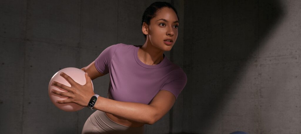 Uma mulher em um ambiente de academia, segurando uma bola de exercício e usando a Xiaomi Smart Band 10 no pulso, demonstrando foco e determinação.