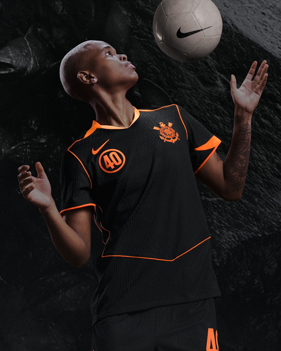 Jogadora usando a nova camiseta do Corinthians, modelo Total 90 da Nike, cor preta com detalhes em laranja, enquanto controla uma bola de futebol.