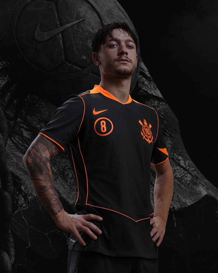 Jogador usando a nova camisa do Corinthians, modelo Total 90, em preto com detalhes em laranja, posicionado em frente a um fundo escuro com elementos gráficos.