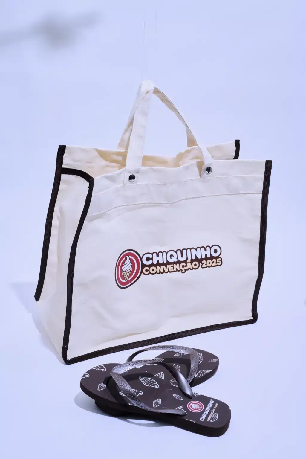 Bolsa de algodão cru com alças reforçadas e logotipo da Chiquinho, acompanhada de chinelos decorados, apresentando o tema da convenção de 2025.