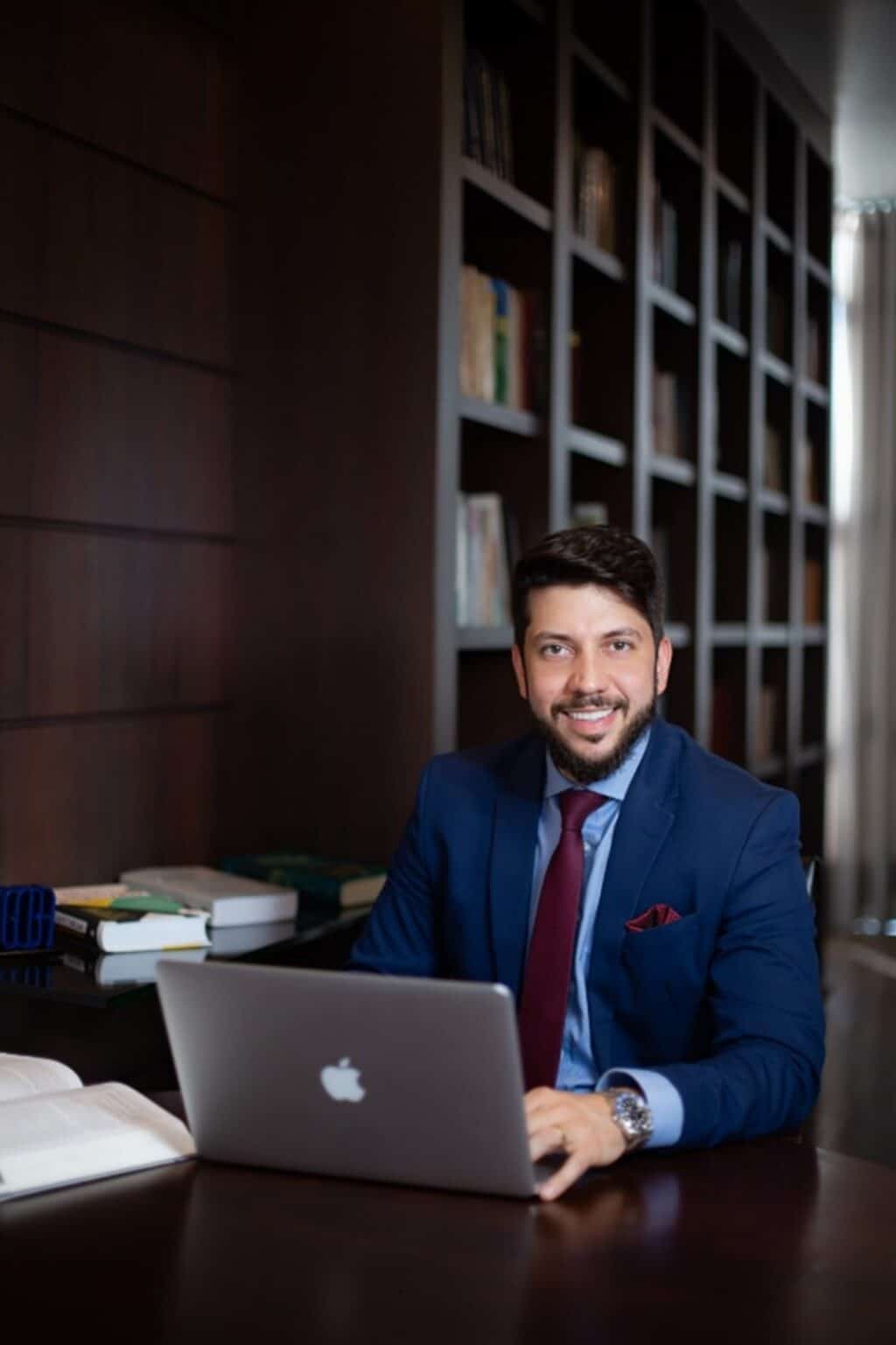 Homem sorridente em um escritório, usando um terno azul e segurando um laptop, com estantes de livros ao fundo.