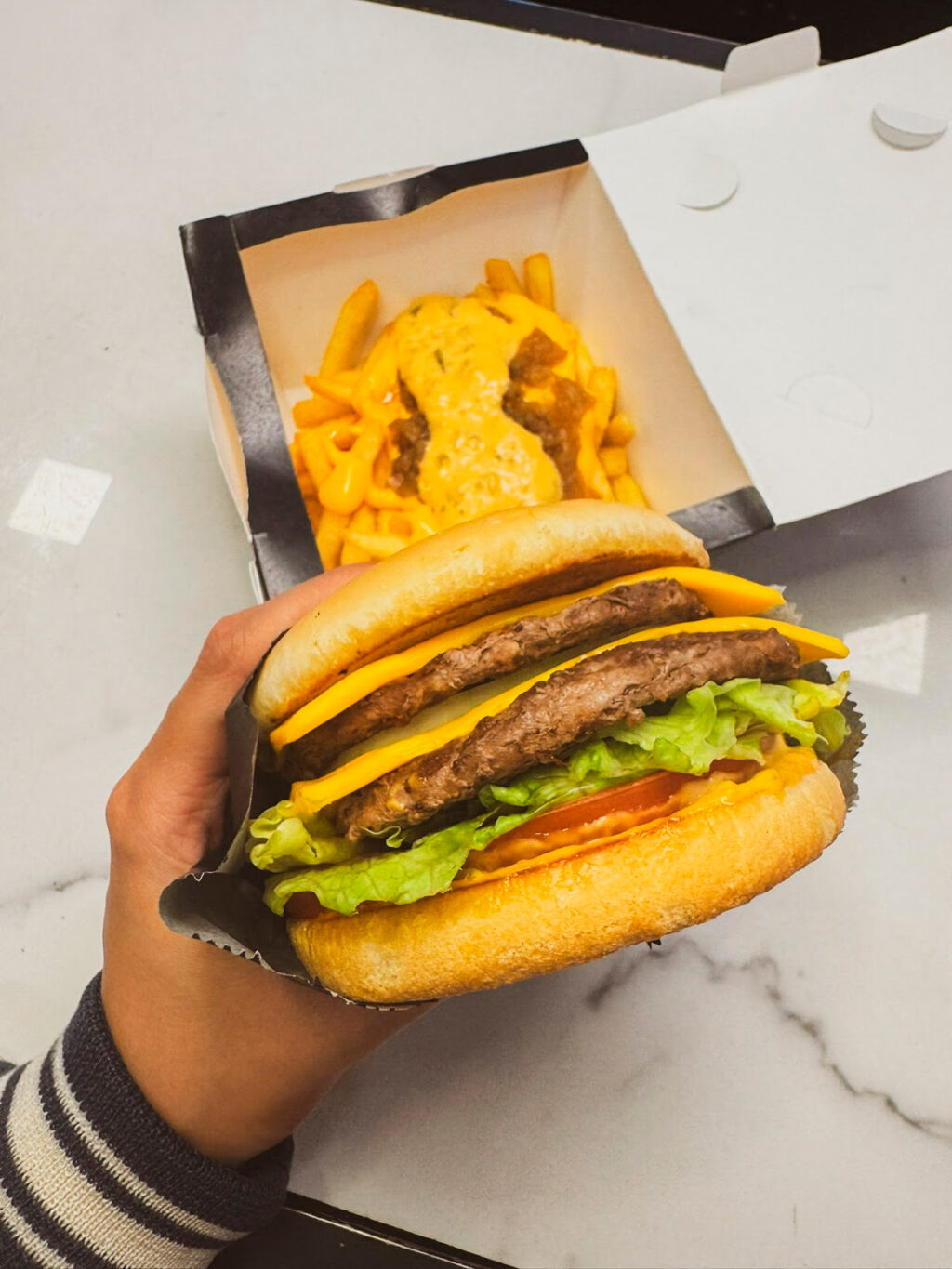 Imagem de um lanche com três hambúrgueres de carne, fatiado de queijo americano, alface e tomate, sendo segurado por uma mão. Ao fundo, uma caixa com batatas fritas cobertas por queijo.