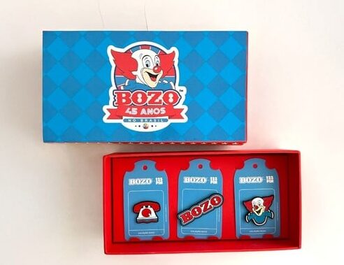Caixa de pins temáticos do personagem Bozo, comemorando 45 anos, com três pins inclusos: um telefone vermelho, o rosto do palhaço e o logo do Bozo.