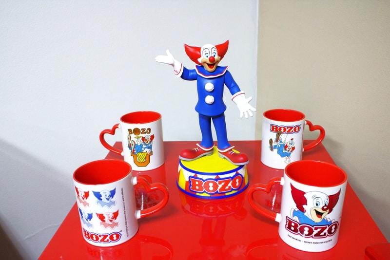 Miniatura colecionável do Palhaço Bozo em pé sobre uma base circular, rodeada por canecas temáticas do personagem, em um fundo colorido.